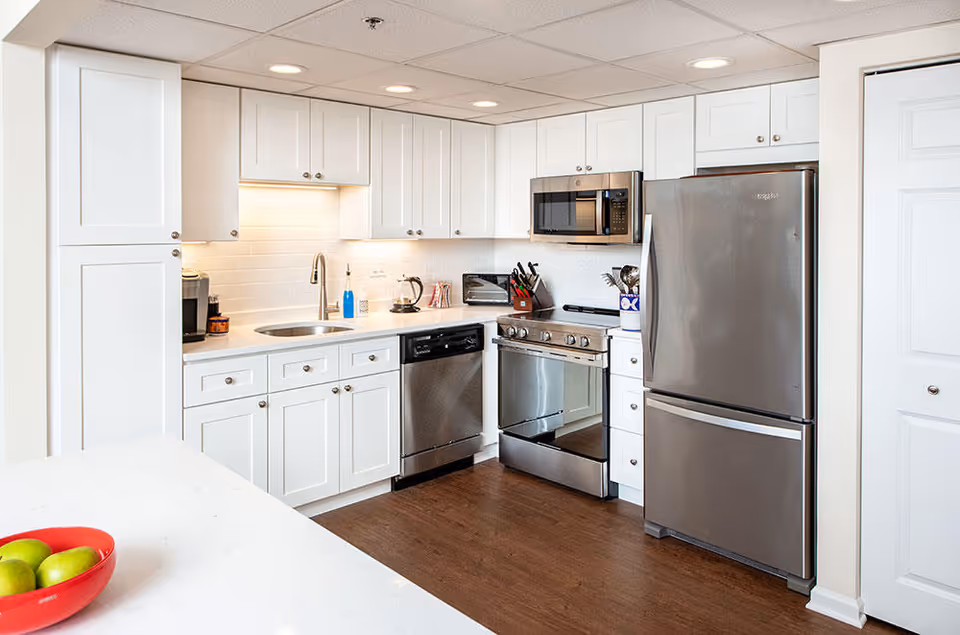 Modern kitchen with white cabinets, stainless steel refrigerator, oven, microwave, and dishwasher. The countertop has a red bowl with green apples. The kitchen has a wooden floor and recessed ceiling lights.