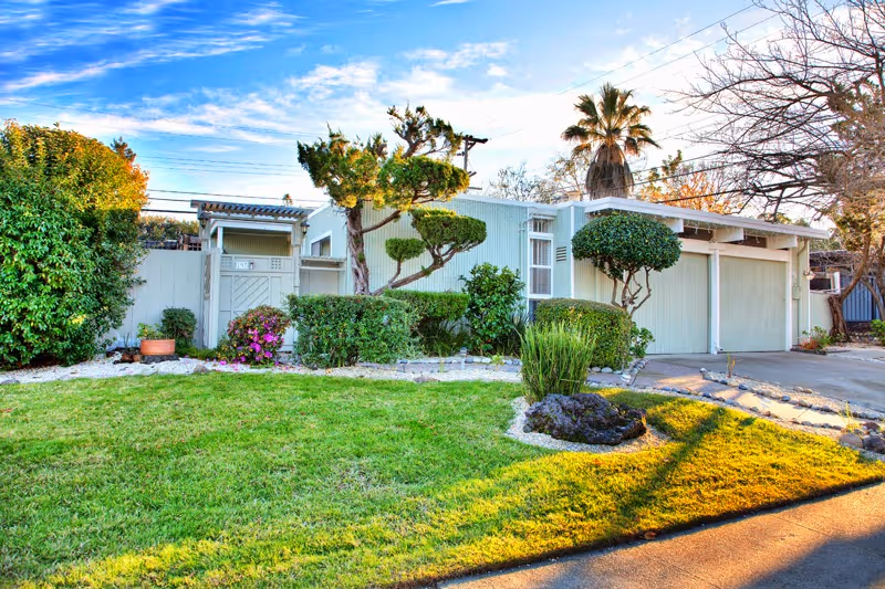 Exterior view of a single-story residential building with a well-maintained front yard featuring green grass, trimmed bushes, and a variety of trees including a palm tree. The building has a light-colored facade with a double garage door and a small entrance gate on the left side.