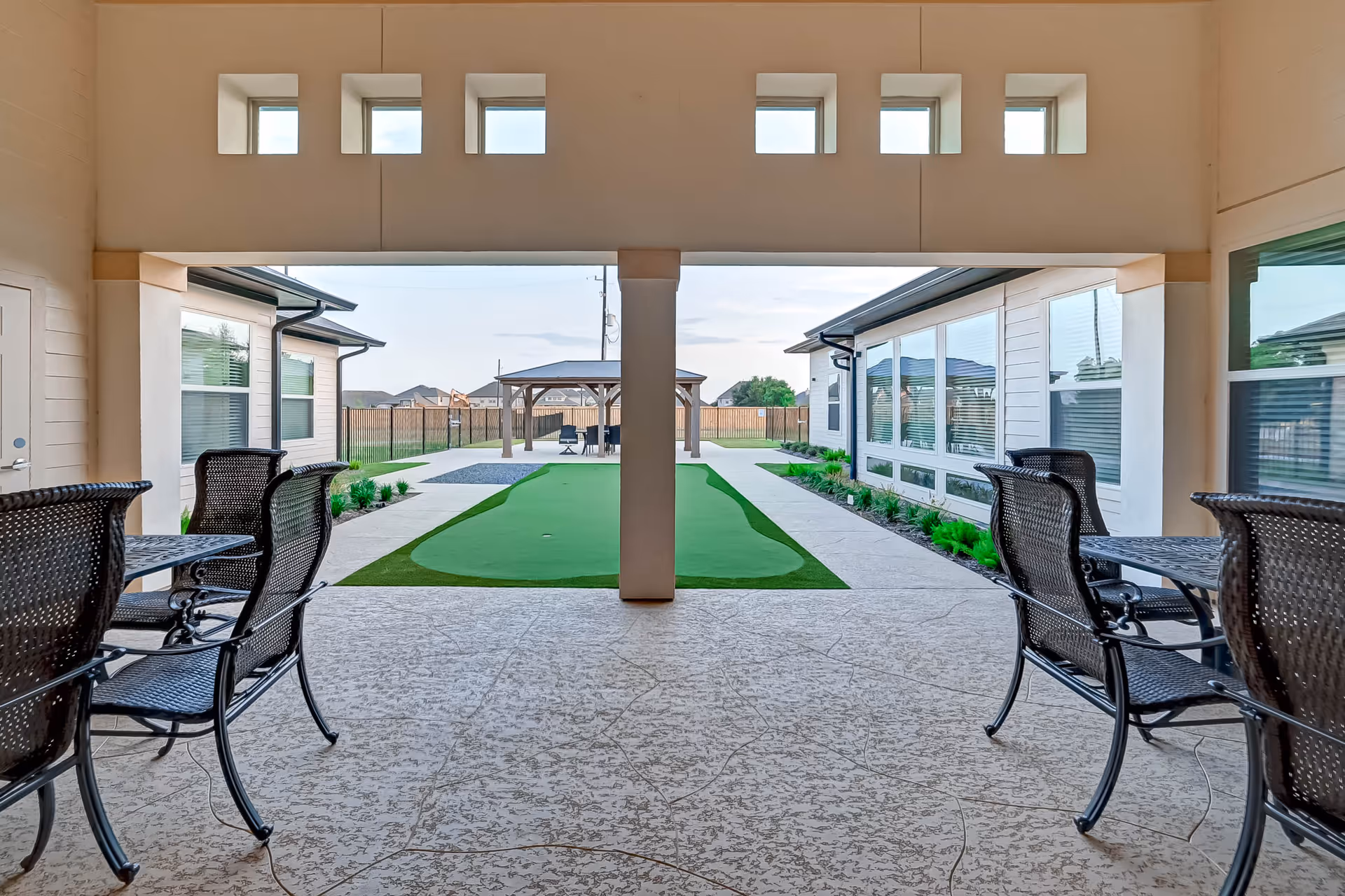 Covered patio area with black metal chairs and tables on a textured concrete floor, overlooking a green artificial putting green and a wooden gazebo in the background, surrounded by a fenced yard and adjacent buildings with large windows.