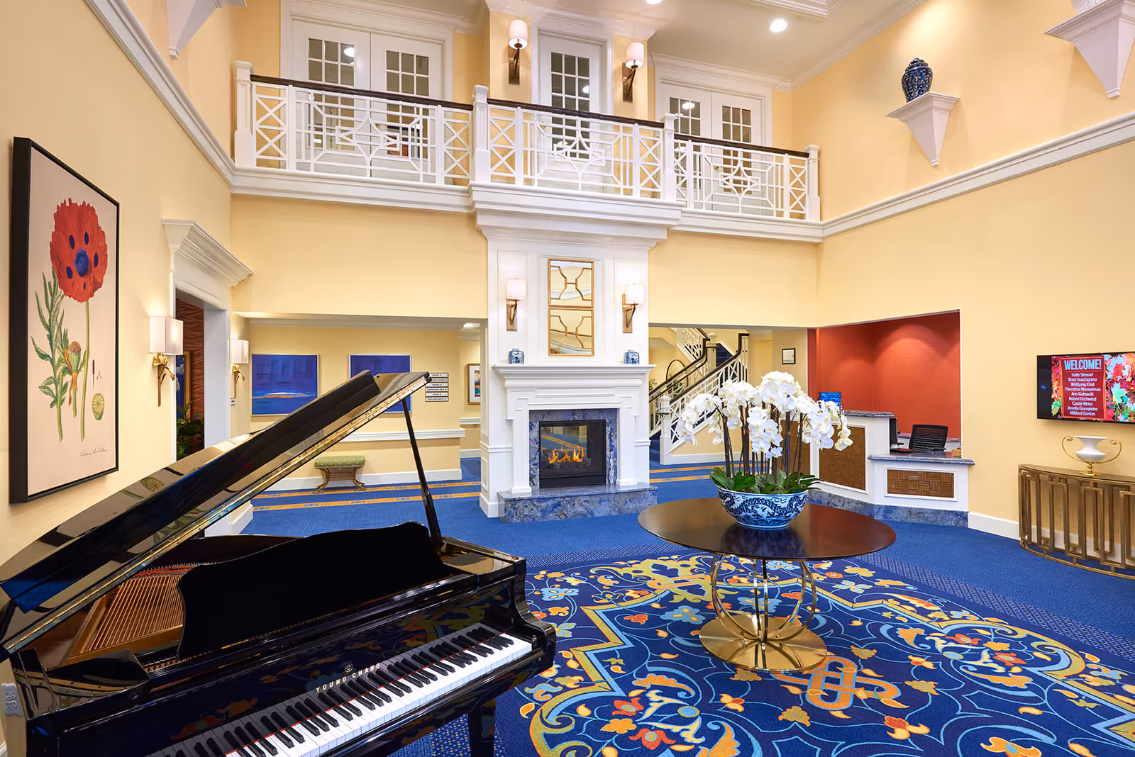 Spacious and elegant assisted living facility lobby with a black grand piano on the left, a round table with a blue and white floral vase holding white orchids in the center, a decorative blue and yellow patterned carpet, a white fireplace with a mirror above it, yellow walls, and a balcony railing on the upper level. There is a reception desk on the right with a red accent wall and a digital welcome sign.