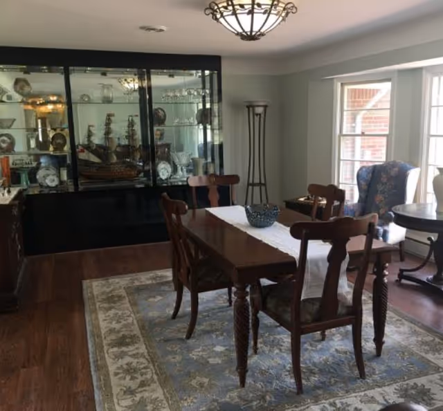 Dining room with a wooden table and chairs on a patterned rug, a glass-front china cabinet, and large windows letting in light.