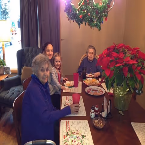 Three elderly women and a young girl sitting around a dining table with holiday-themed placemats, enjoying a meal together. A large poinsettia plant and a festive wreath hanging above the table are visible in the room.