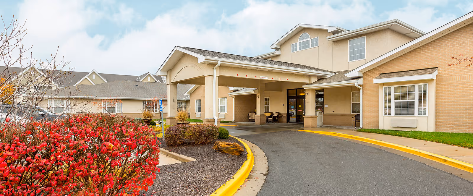 Exterior view of the entrance to Brookdale Rosehill senior living facility with a covered drop-off area, beige buildings, and red bushes in the foreground under a partly cloudy sky.