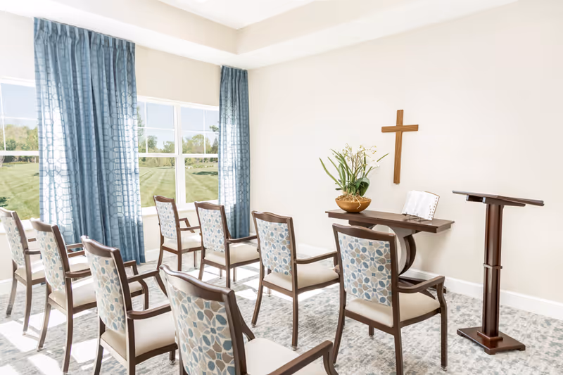 A small chapel or prayer room with wooden chairs arranged in rows facing a wooden table with a flower arrangement and an open book. A wooden cross is mounted on the wall above the table. Large windows with blue patterned curtains let in natural light, and the room has light-colored walls and carpeted flooring.