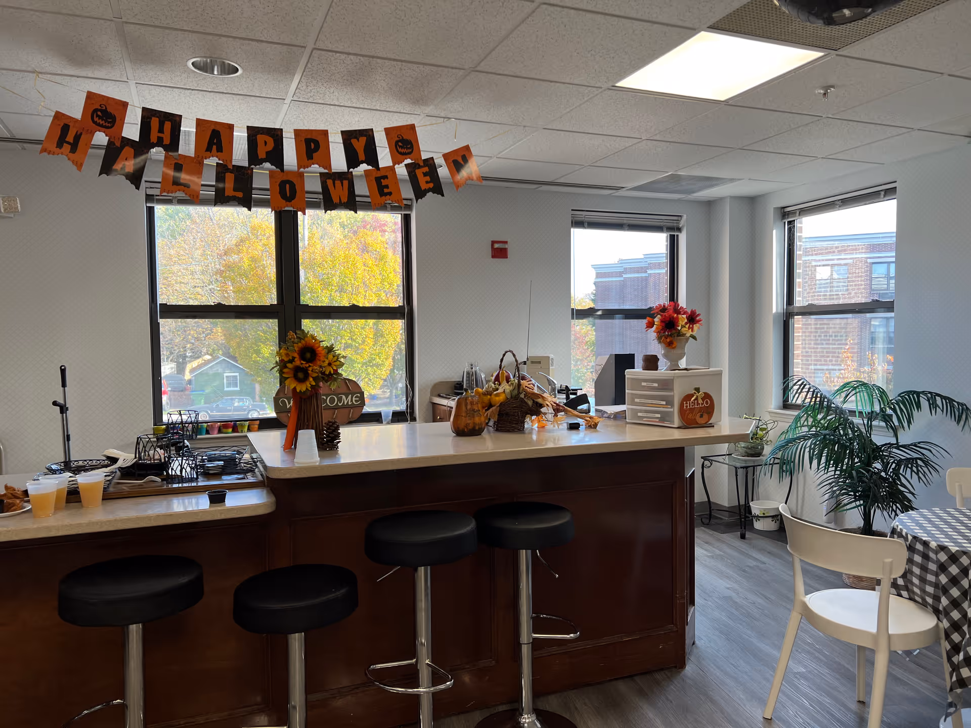Interior view of a communal dining or kitchen area decorated for Halloween with a 'Happy Halloween' banner hanging above a counter. The counter has stools, fall-themed decorations including flowers, pumpkins, and a basket. Large windows show trees with autumn foliage and a brick building outside. A small table with a checkered tablecloth and white chairs is visible on the right side along with a potted plant.