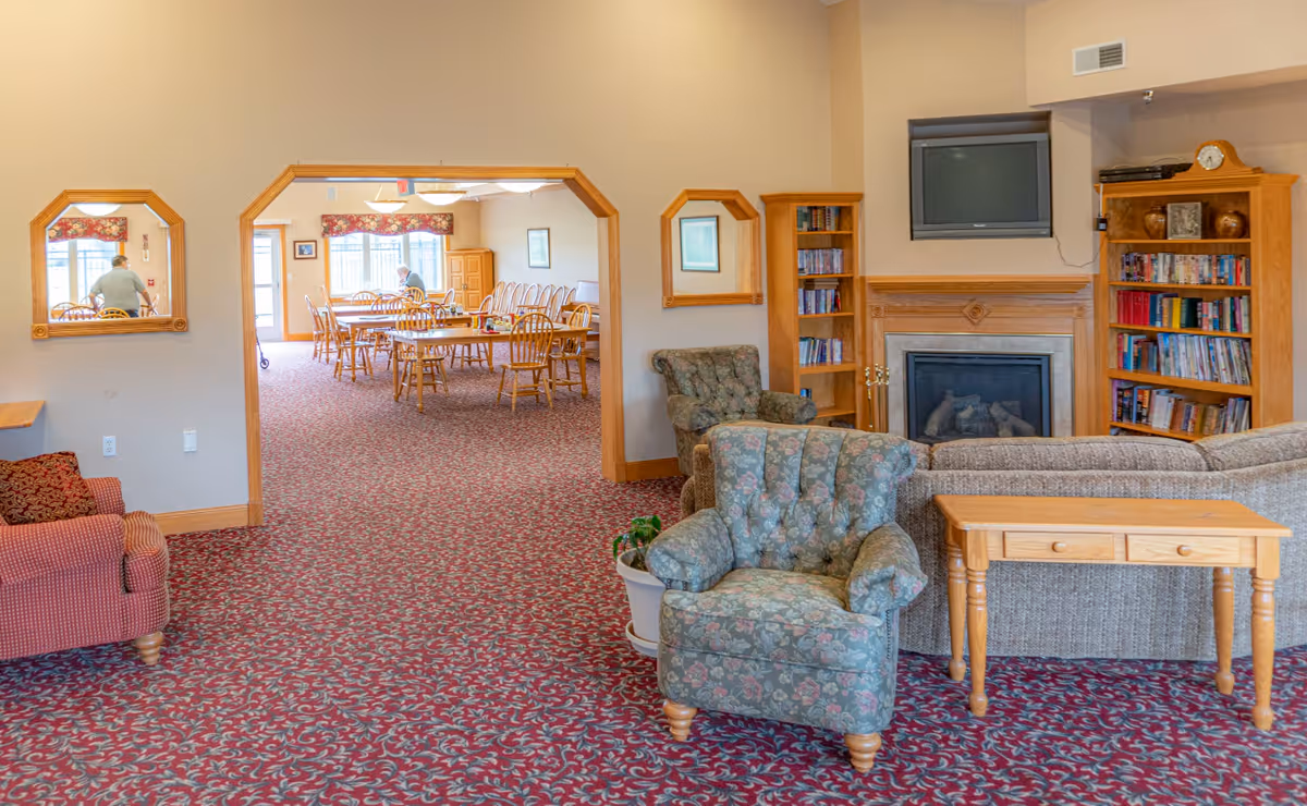 Interior view of a senior living facility showing a cozy common area with floral upholstered armchairs, a sofa, wooden bookshelves filled with books, and a fireplace with a TV mounted above it. Through a large open doorway, a dining area with wooden tables and chairs is visible, with two elderly individuals seated at the tables. The room has patterned carpet and light-colored walls with wooden trim.