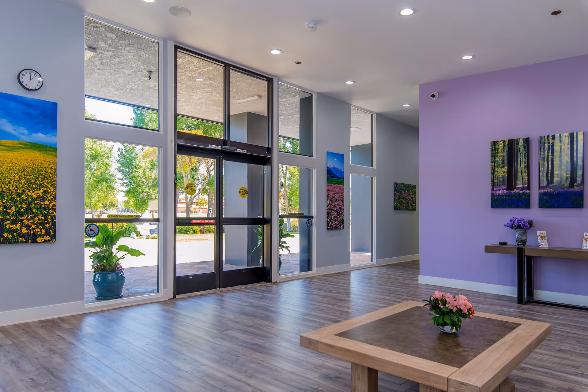 Spacious assisted living lobby with glass entrance doors, a central table with flowers, purple accent wall and nature artwork.