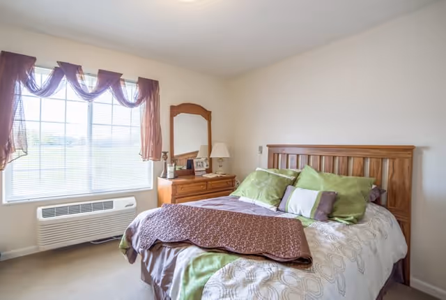 A bright bedroom with a large window covered by sheer purple curtains. The room features a wooden bed with green and white bedding, a brown blanket folded at the foot of the bed, and a wooden dresser with a mirror, lamp, and framed photos.
