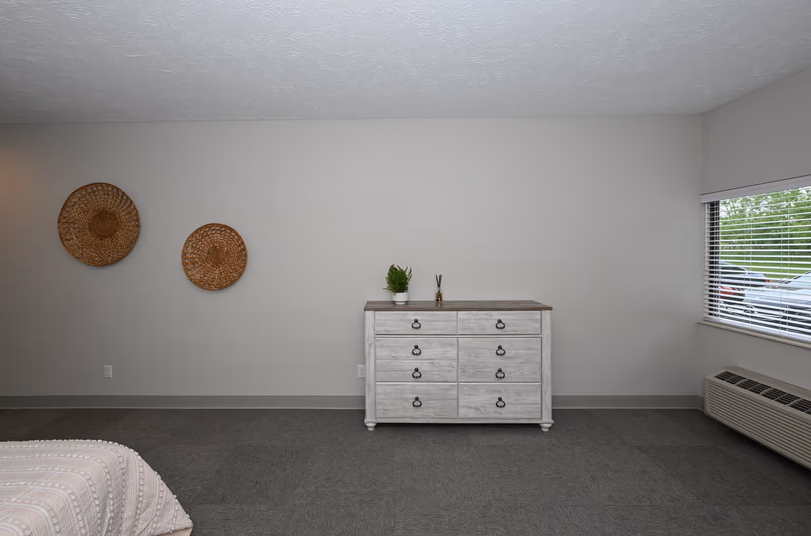 Minimal bedroom interior showing a white wooden dresser against a pale gray wall, two woven wall baskets, a window with blinds, and a partial bed in the corner.