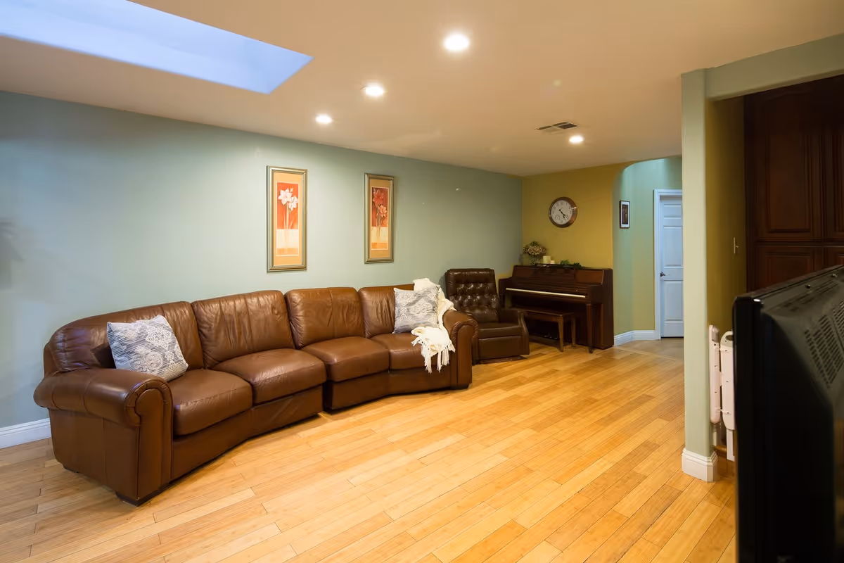 A cozy living room with a brown leather sectional sofa adorned with two patterned pillows and a white throw blanket. There is a brown leather armchair next to a wooden piano with a bench. The walls are painted light green and yellow, decorated with two framed floral paintings and a round clock. The floor is wooden, and recessed ceiling lights illuminate the room. A television is partially visible in the foreground.