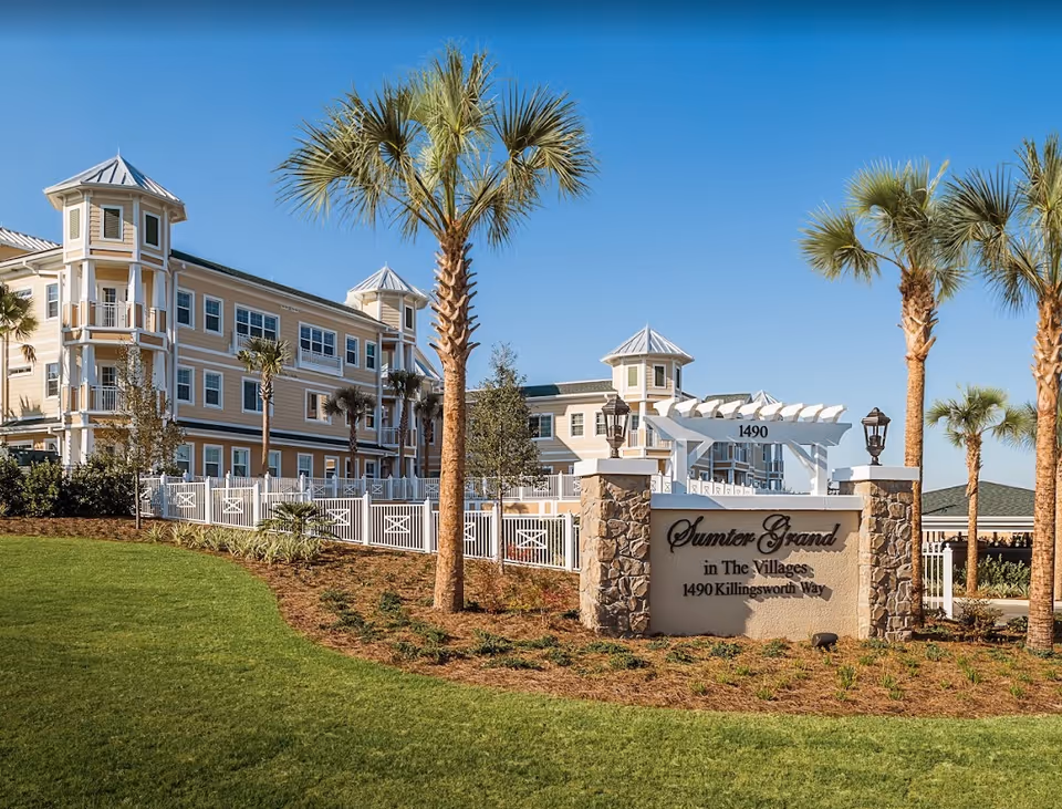 Exterior view of Sumter Grand in The Villages, a multi-story senior living facility with beige siding, white trim, and multiple small towers with metal roofs. The building is surrounded by palm trees, a white fence, and landscaped greenery under a clear blue sky. A stone and stucco sign in the foreground displays the facility name and address: 1490 Killingsworth Way.