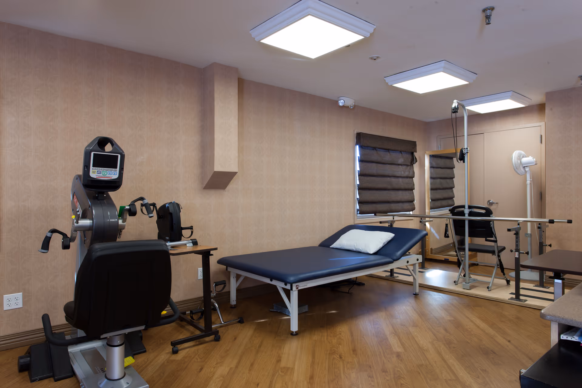 A rehabilitation room with a treatment table, exercise bike, parallel bars and other therapy equipment on wood flooring.
