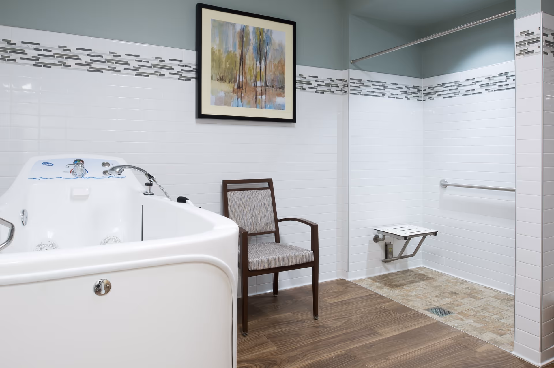 A clean and modern bathroom featuring a white walk-in bathtub with a faucet and controls, a wooden chair with a cushioned seat, a wall-mounted foldable shower seat, and a grab bar. The walls are tiled with white tiles and a decorative horizontal strip of smaller tiles. A framed painting of a nature scene hangs on the wall above the chair. The floor has wood-like and tiled sections.