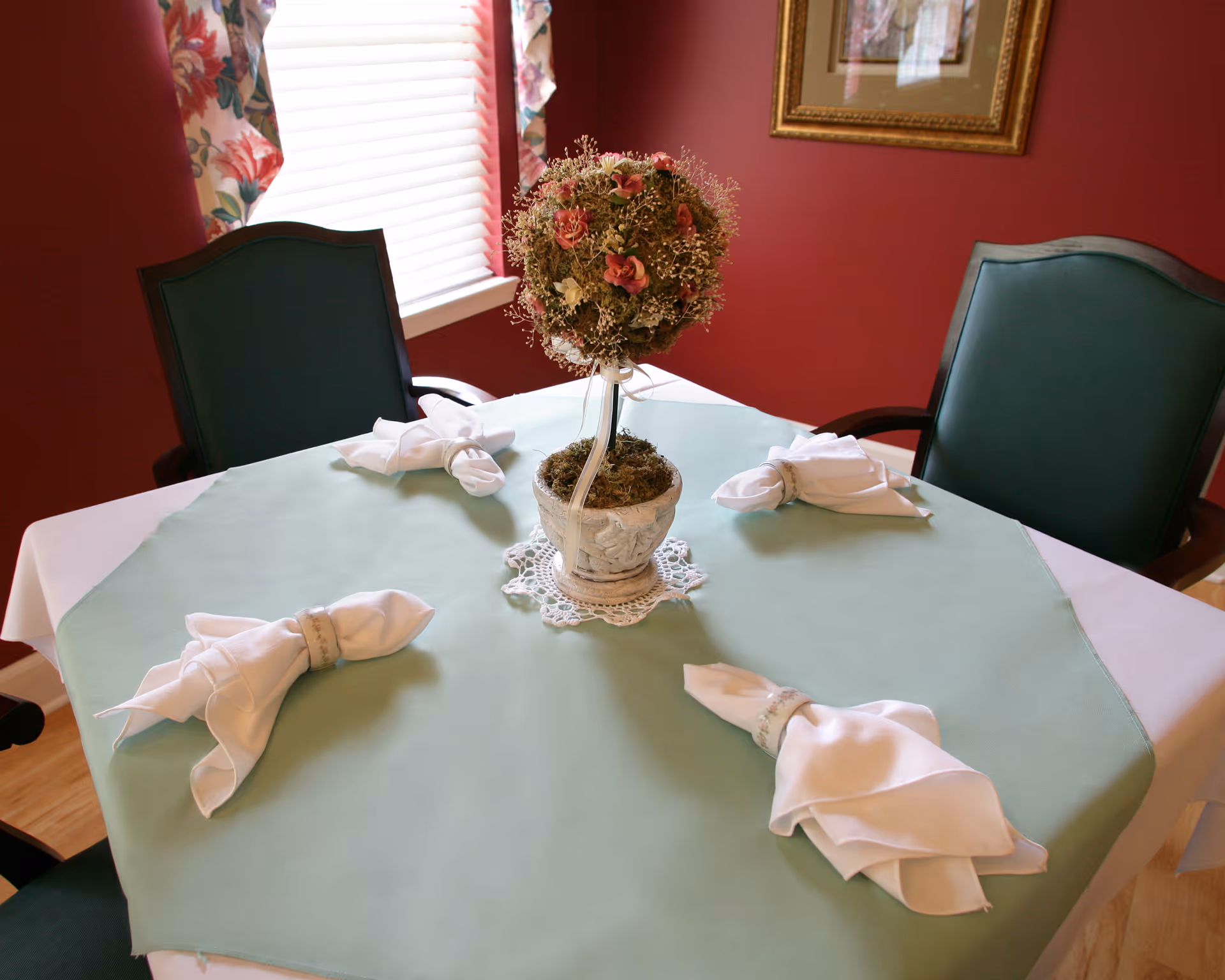 A dining table set with a green tablecloth and white napkins folded elegantly with napkin rings. A decorative floral centerpiece in a pot is placed in the middle of the table. Two green cushioned chairs are visible, with a window and floral curtains in the background and a framed picture on the red wall.