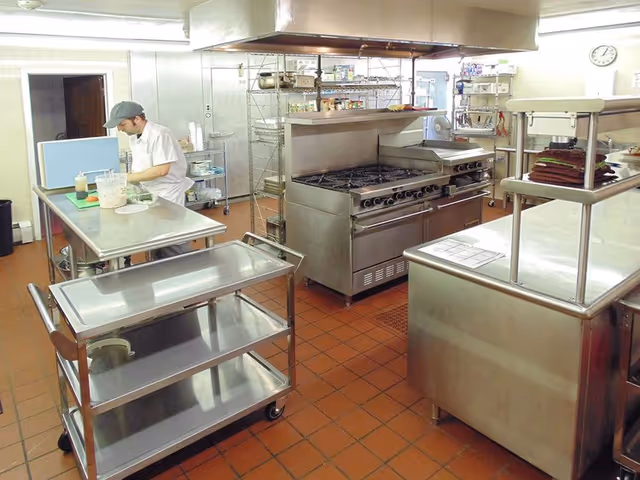 A commercial kitchen with stainless steel appliances and work surfaces. A person wearing a white uniform and a gray cap is preparing food on a counter. The kitchen has a large stove with multiple burners, shelves with various ingredients, and a stainless steel cart with trays. The floor is covered with reddish-brown tiles.