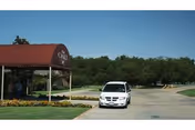 Entrance area of The Oaks of Granbury facility with a maroon canopy displaying the facility name, a white car parked on the driveway, and trees in the background under a clear blue sky.