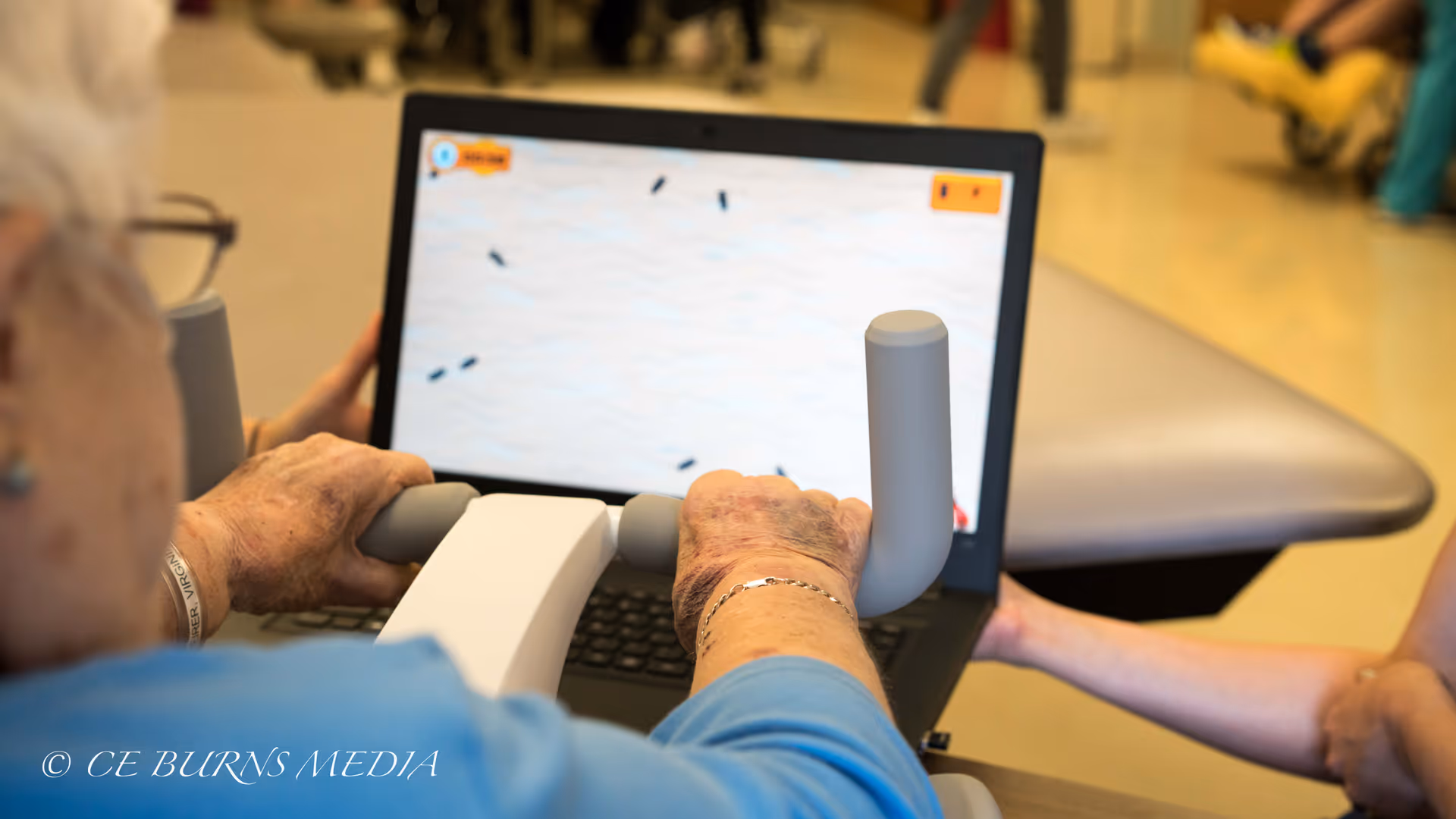 An elderly person using a hand exercise device connected to a laptop displaying a game or interactive activity, with another person assisting by holding the device.