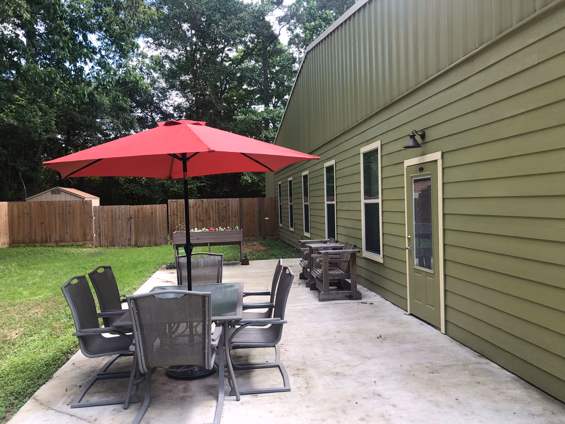 Outdoor patio area with a green building wall on the right side, featuring a door and several windows. There is a concrete patio with a glass table and six chairs under a large red umbrella. Two wooden benches are placed against the building wall. The patio is adjacent to a grassy yard with a wooden fence and trees in the background.
