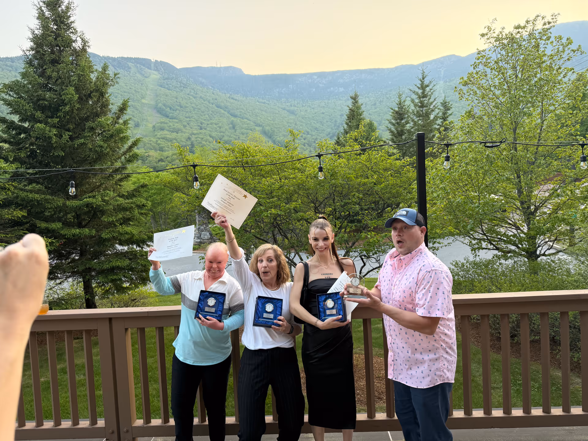 Four people standing on a wooden deck outdoors with a scenic mountain and forest background. Three of them are holding plaques and certificates, celebrating an achievement. One person is taking a photo of the group.