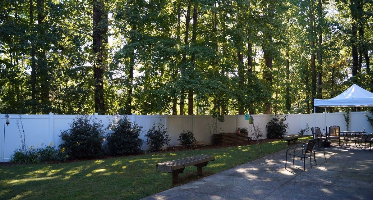 Sunlit fenced backyard with patio seating, benches, a canopy and tall trees beyond a white privacy fence.