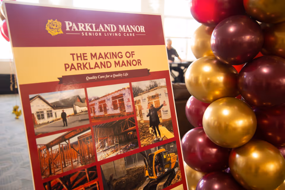 A display board titled 'The Making of Parkland Manor' showing photos of the construction process of Parkland Manor senior living care facility. The board is next to a column of maroon and gold balloons, with a blurred background of an indoor space.