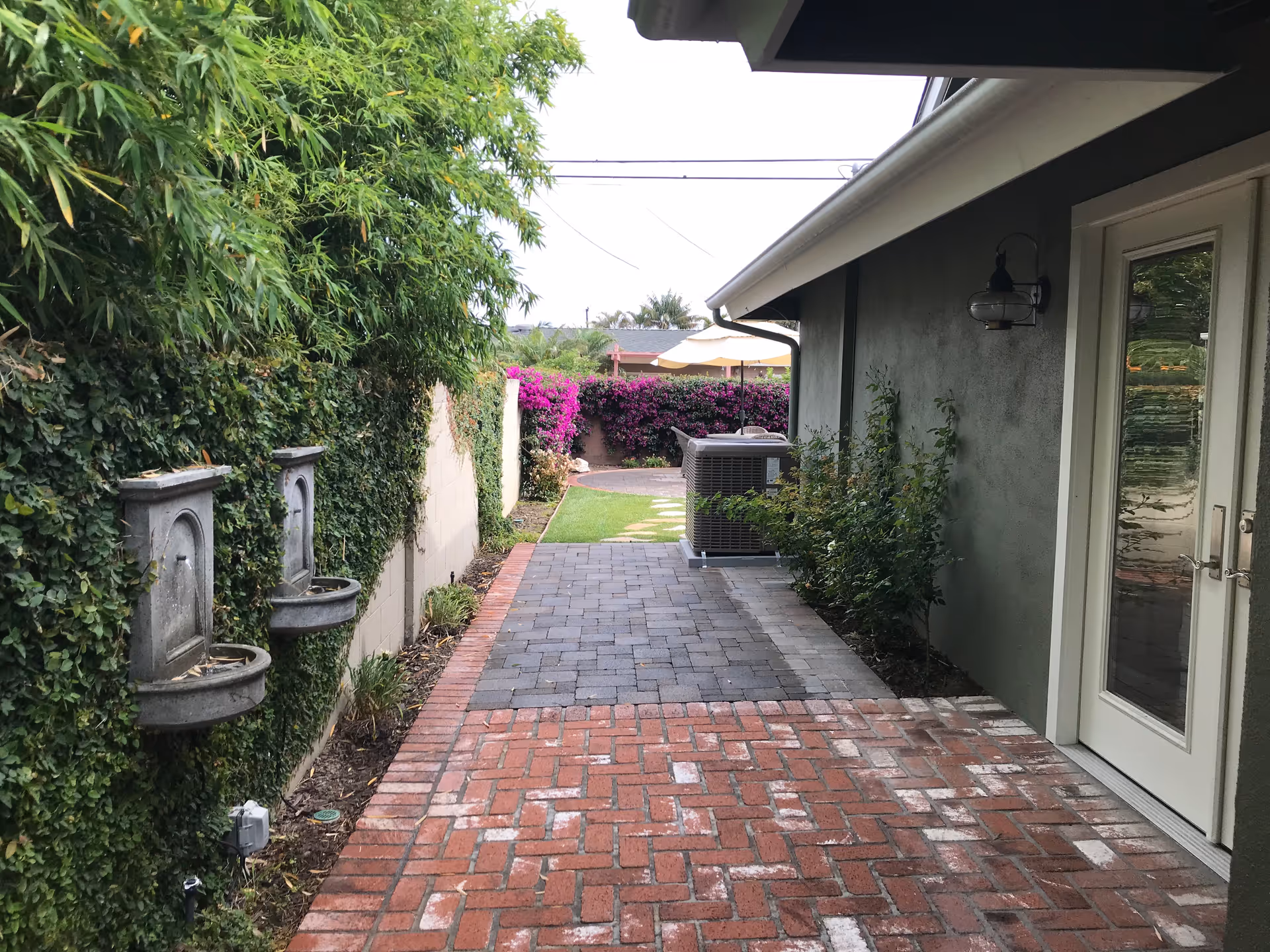 A narrow outdoor walkway beside a building with a green wall on the left covered in ivy and two wall-mounted water fountains. The walkway is paved with red bricks and gray stones leading to a grassy area with purple flowering bushes and an outdoor umbrella in the background. The right side shows the exterior wall of a building with a glass door and a wall-mounted light fixture.