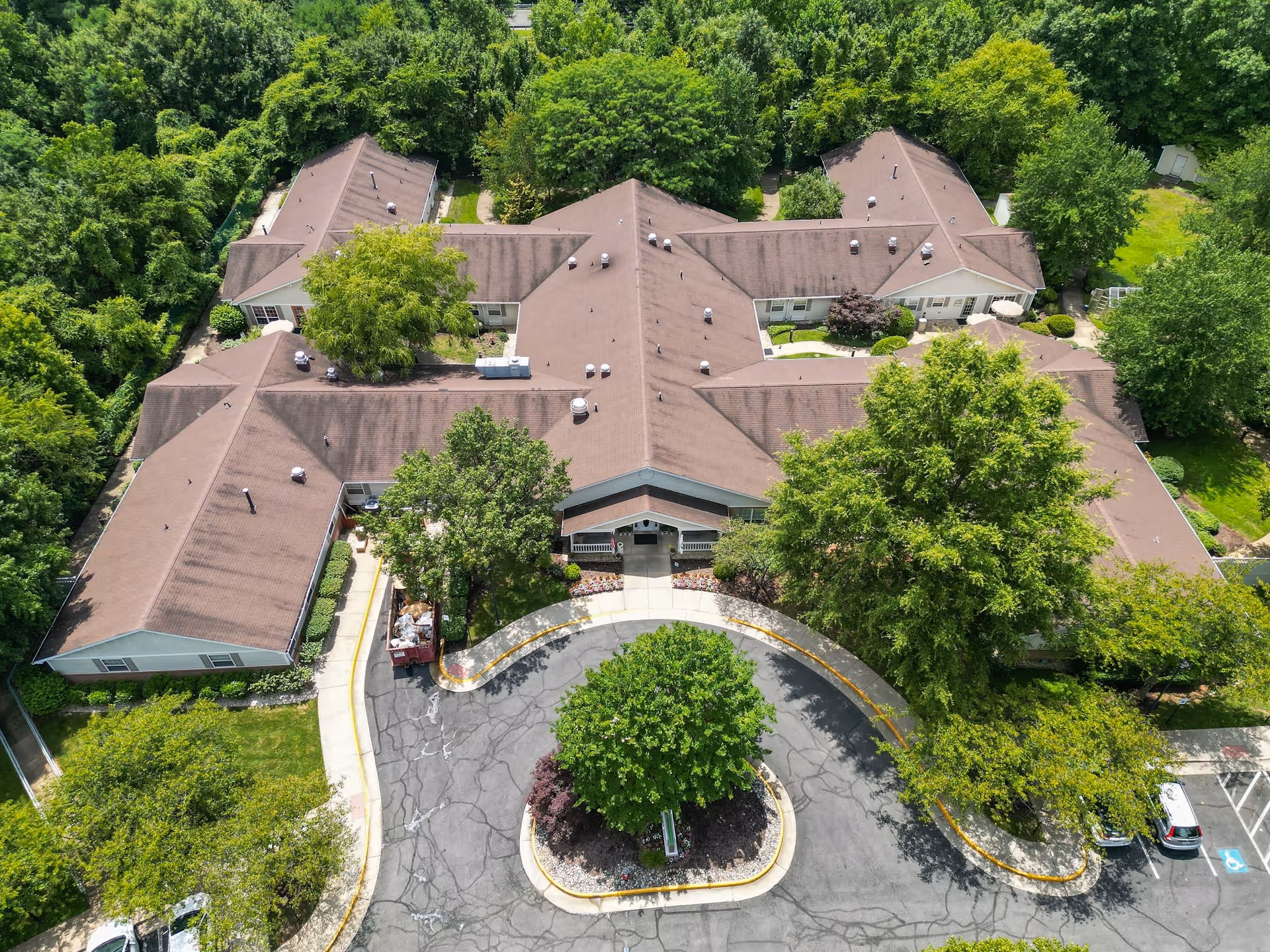 Aerial view of Arden Courts - ProMedica Memory Care Community (Fair Oaks), showing a large, single-story building with a brown roof surrounded by lush green trees and landscaping. The building has a circular driveway with a landscaped island in the center, and several parked cars are visible in the parking lot.