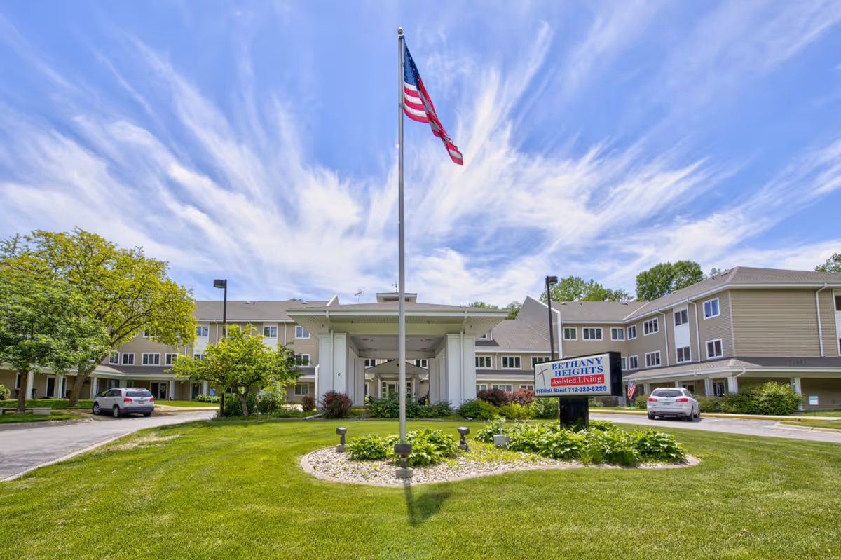 Front exterior of a multi-story assisted living facility with a central covered entrance, an American flag on a tall flagpole, a sign reading 'Bethany Heights Assisted Living', landscaped lawn and parked cars.