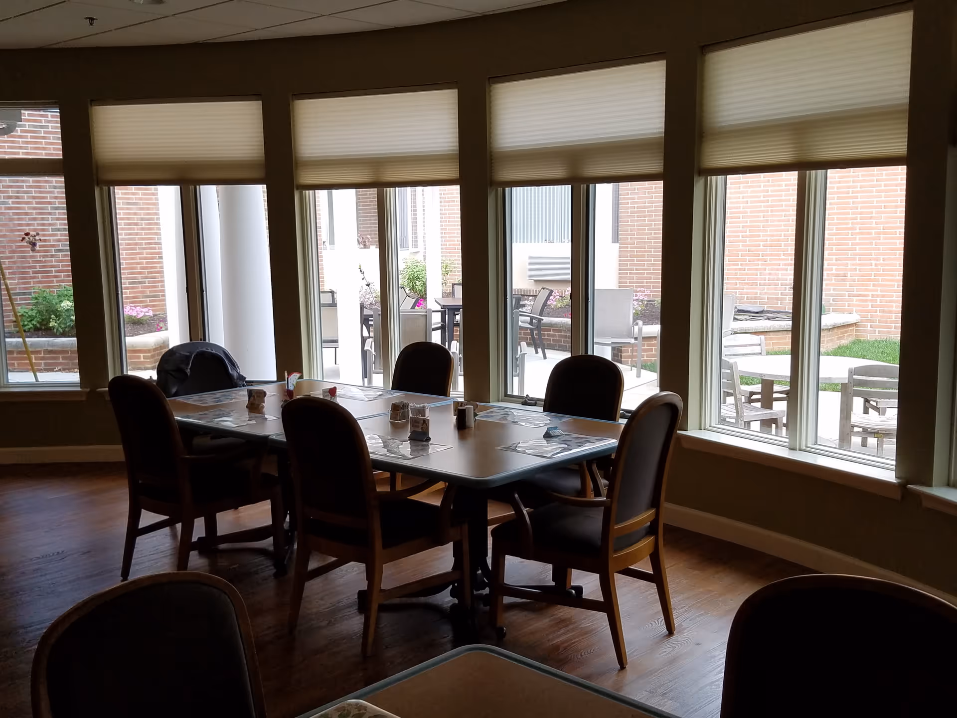 A dining area with a rectangular table surrounded by six chairs. The table has placemats and small condiment containers on it. Large windows with partially lowered blinds provide a view of an outdoor patio area with additional tables and chairs, brick walls, and some greenery.