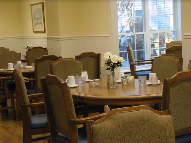 A dining room with multiple wooden tables and cushioned chairs arranged around them. Each table has white cups, glasses, and small flower vases with white flowers. There is a large window letting in natural light and a framed picture on the wall.