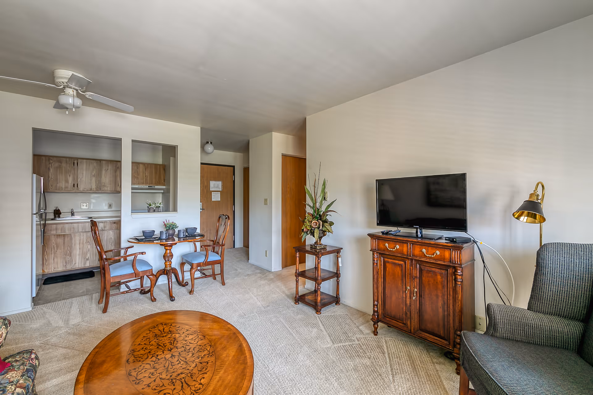 A cozy living room and dining area in a senior living facility. The room features a wooden coffee table with intricate designs, a dark upholstered armchair, a wooden cabinet with a flat-screen TV on top, and a floor lamp with a gold shade. There is a small wooden dining table set with two chairs and place settings. The kitchen area with wooden cabinets and a refrigerator is visible through an open wall space. The walls are light-colored, and the floor is carpeted.