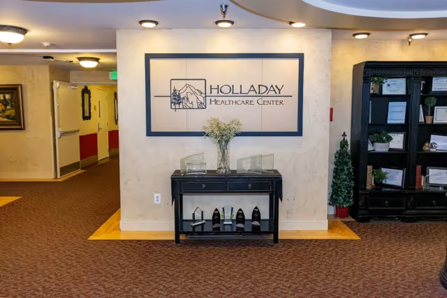 Interior view of Holladay Healthcare Center featuring a beige wall with the facility's logo mounted above a black console table displaying several awards and a vase with flowers. To the right is a black bookshelf with framed certificates and decorative plants. The floor is carpeted in a patterned design, and there is a hallway visible to the left.