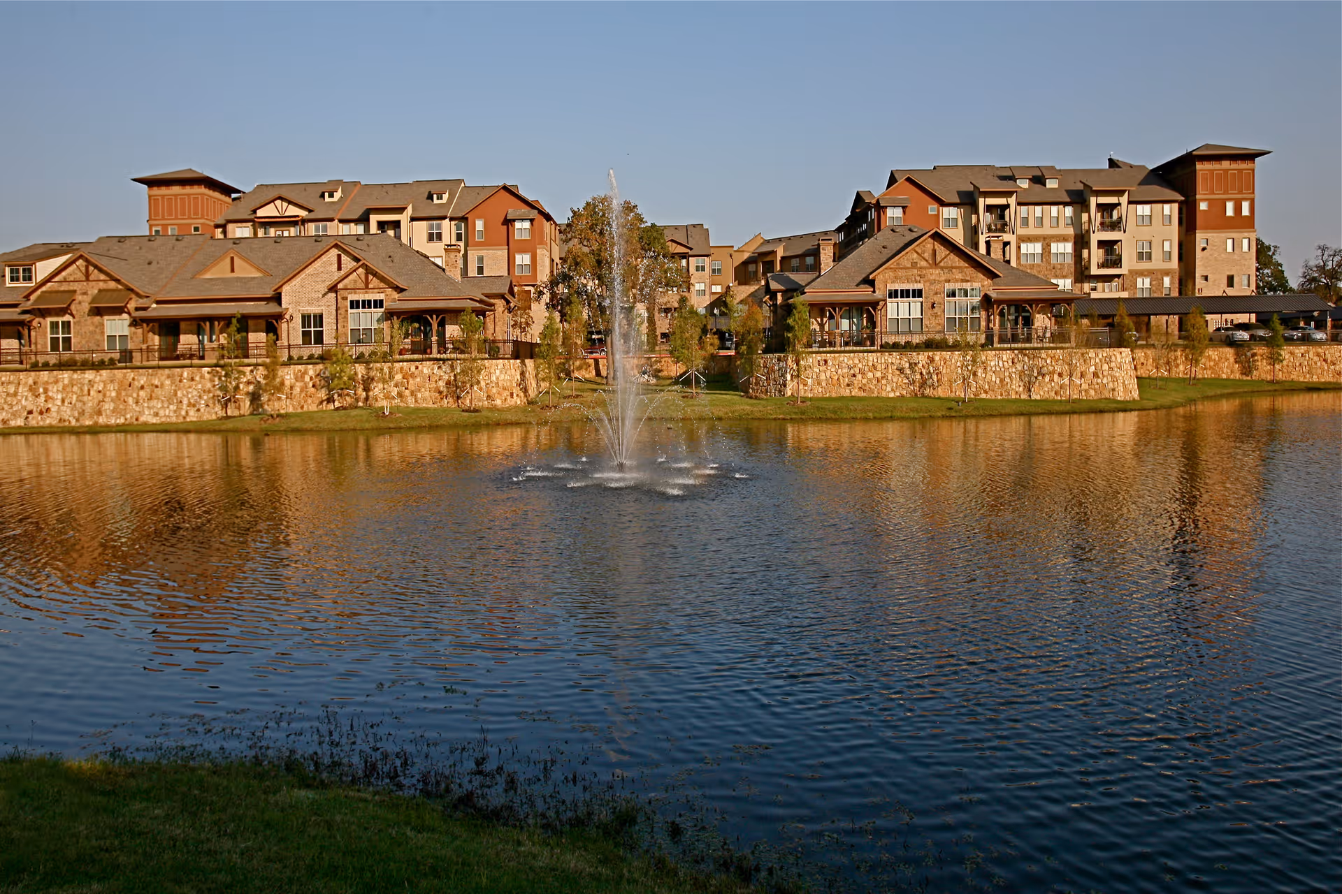 View of a senior living facility named Watercrest at Mansfield with multiple buildings featuring stone and wood exteriors, situated behind a large pond with a central water fountain.