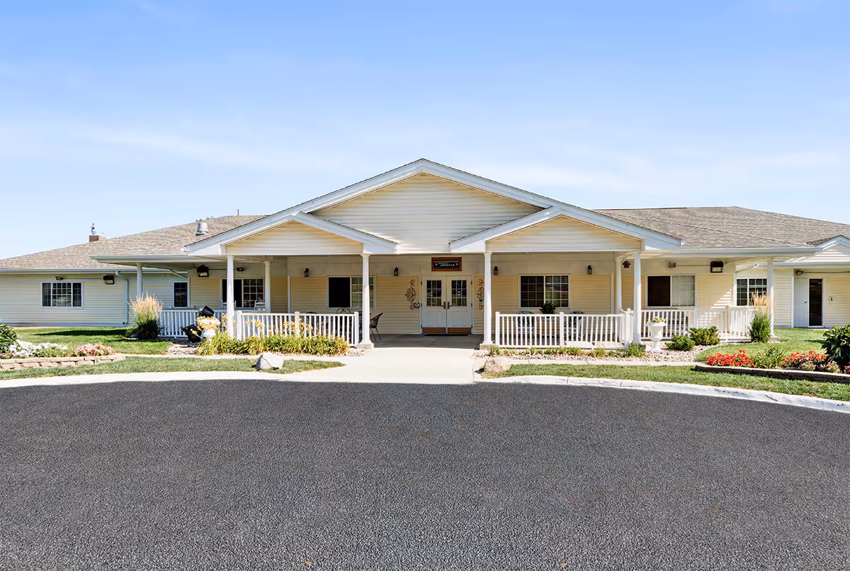 Front exterior view of Carter Place Senior Living, a single-story building with a covered entrance, white railings, and landscaped garden beds with flowers and shrubs. The sky is clear and blue.