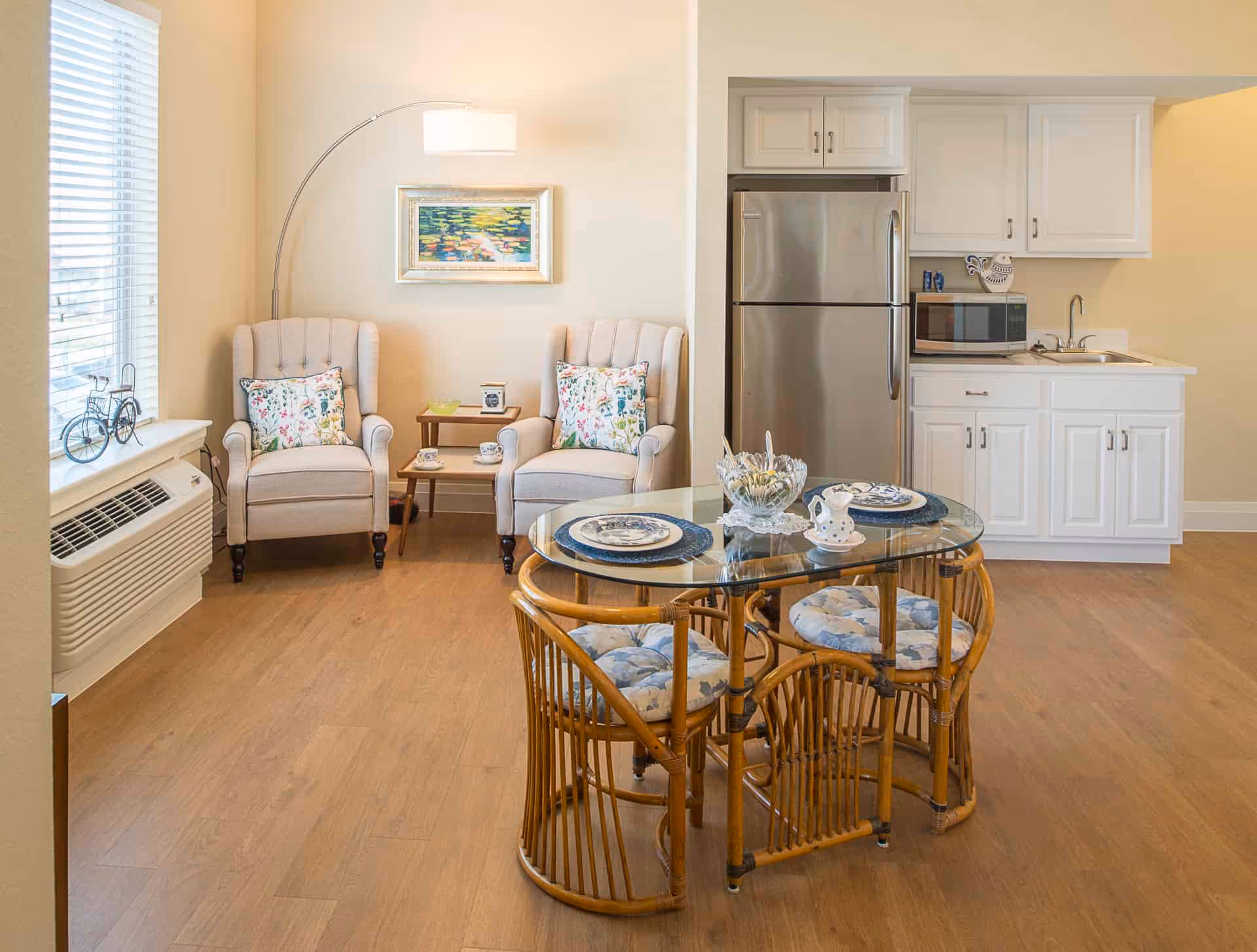 A cozy living and dining area featuring two beige armchairs with floral cushions, a small wooden side table with a clock and cups, a glass-top dining table with four rattan chairs, and a compact kitchen area with white cabinets, a stainless steel refrigerator, a microwave, and a sink.