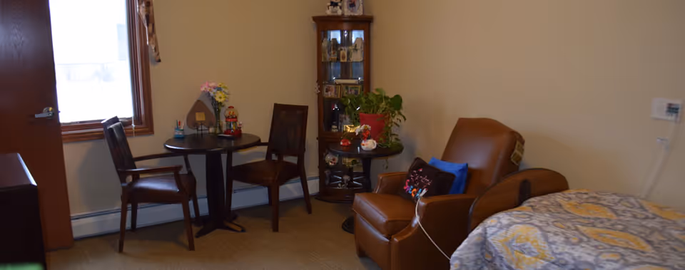A cozy room with a small round table and two chairs near a window, a brown leather armchair with pillows, a bed with a patterned blanket, and a corner cabinet displaying various decorative items and plants.