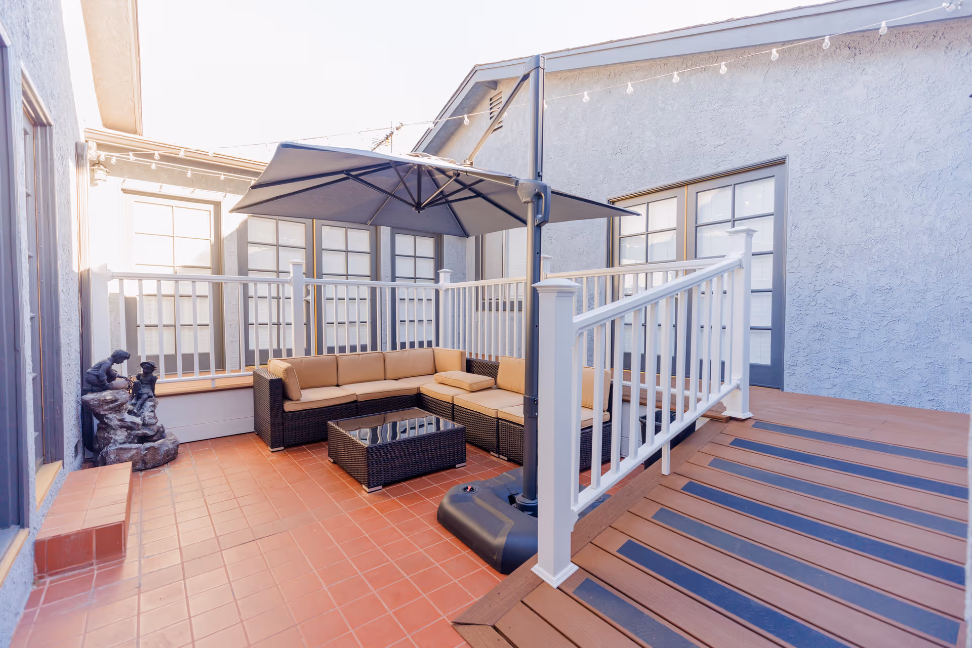 Outdoor patio area with terracotta tile flooring, a brown wicker sectional sofa with beige cushions, a glass-top coffee table, a large gray umbrella providing shade, white railing, and a small statue of two children on the left side near the wall.