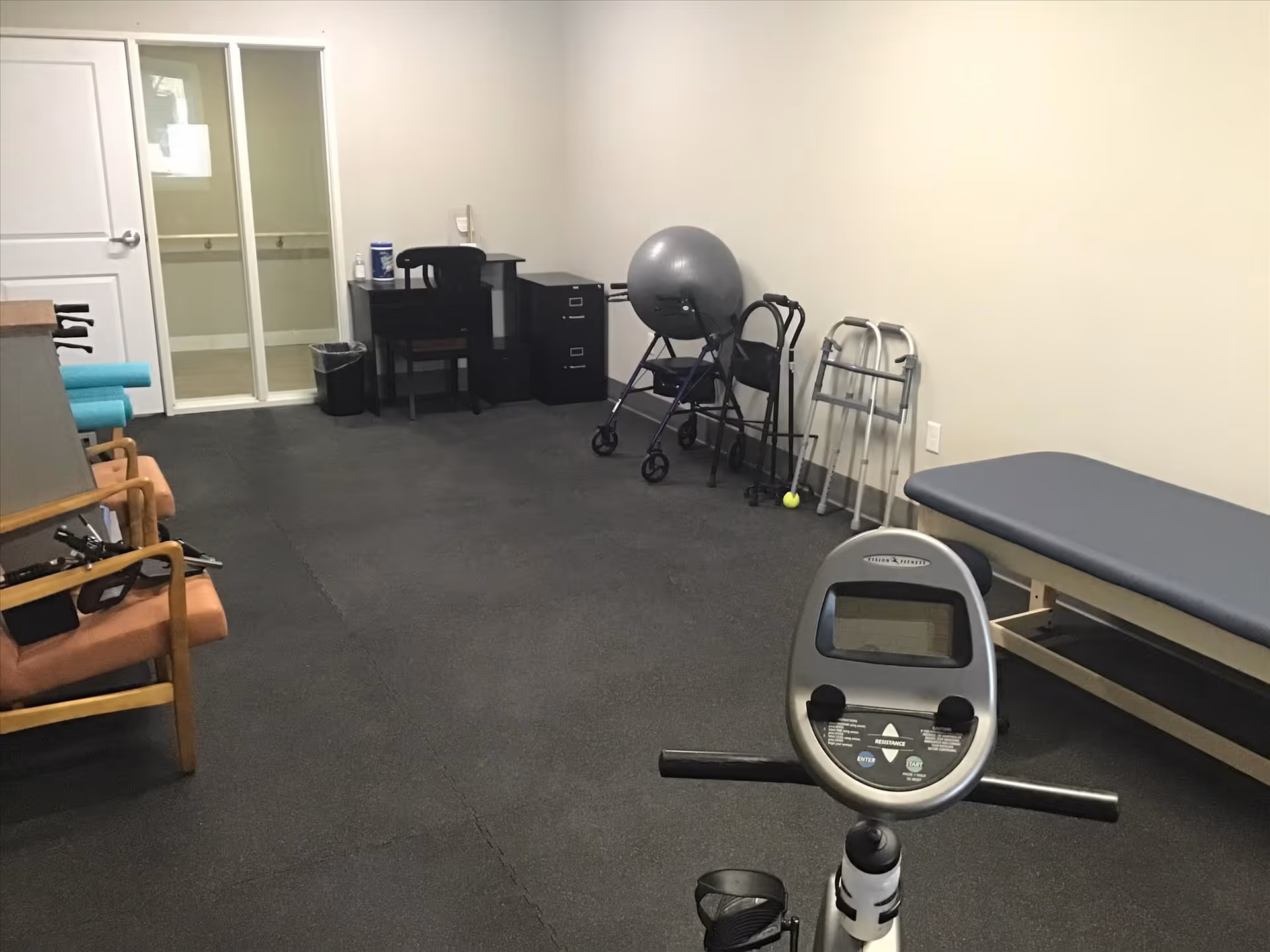 A small exercise and therapy room with a stationary exercise bike in the foreground, a padded therapy table on the right, several walkers and a large exercise ball against the back wall, a desk with a chair and filing cabinets, and two wooden chairs with armrests on the left side.
