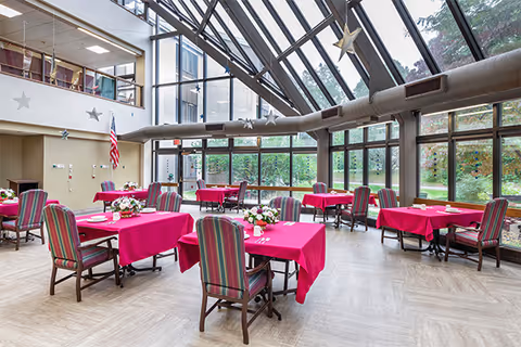 A bright dining room with large glass windows and a glass ceiling letting in natural light. The room has several tables covered with pink tablecloths, each surrounded by striped upholstered chairs. Floral centerpieces decorate the tables. An American flag is displayed near the wall, and greenery is visible outside through the windows.