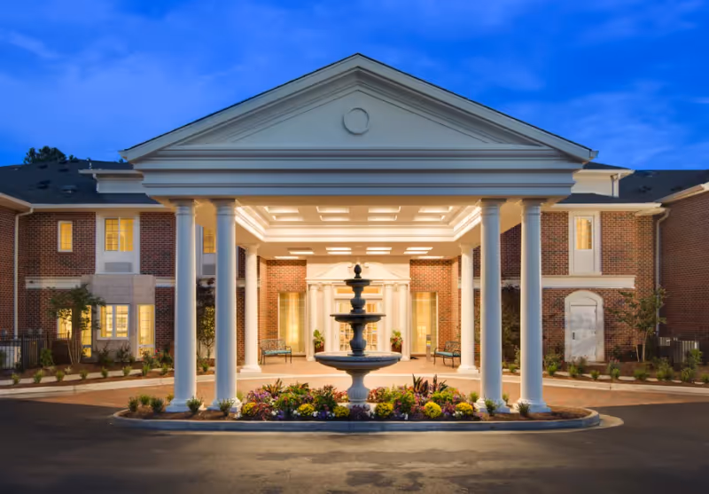 Front exterior view of The Georgian Lakeside facility at dusk, featuring a grand entrance with white columns, a three-tiered fountain surrounded by flowers, and warm lighting illuminating the brick building.