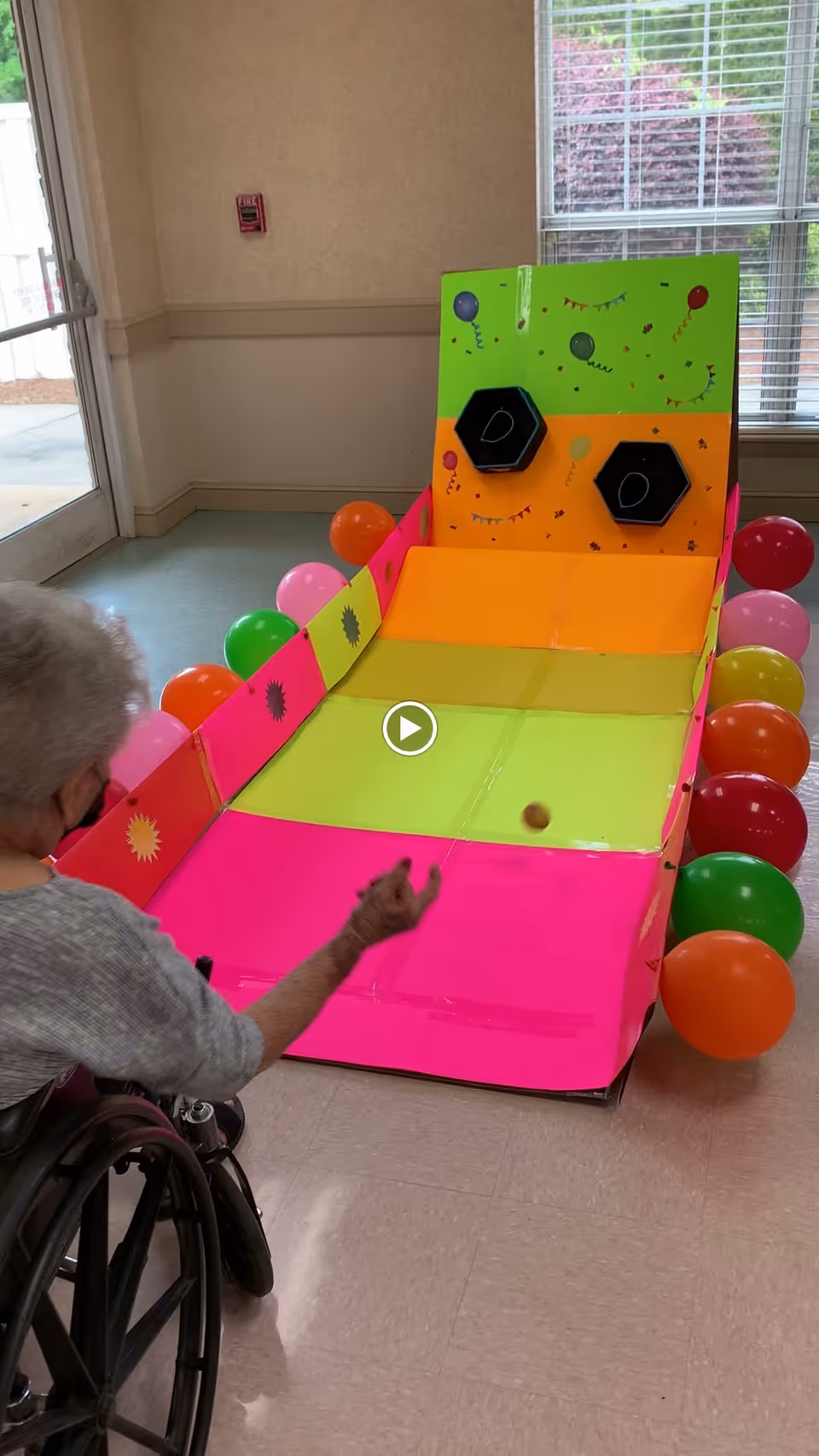 An elderly person in a wheelchair playing a colorful indoor carnival-style ball toss game with hexagonal holes labeled with the letter D, surrounded by balloons in a well-lit room with a window and a door.