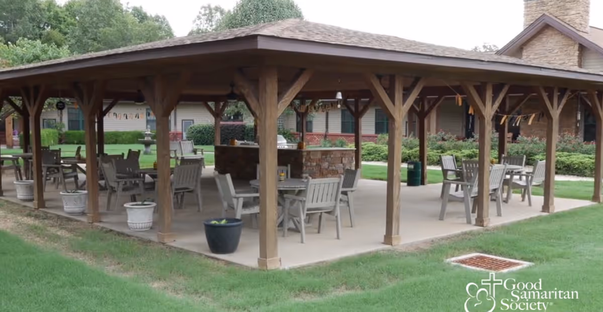 Wooden covered outdoor pavilion with tables and chairs on landscaped grounds.