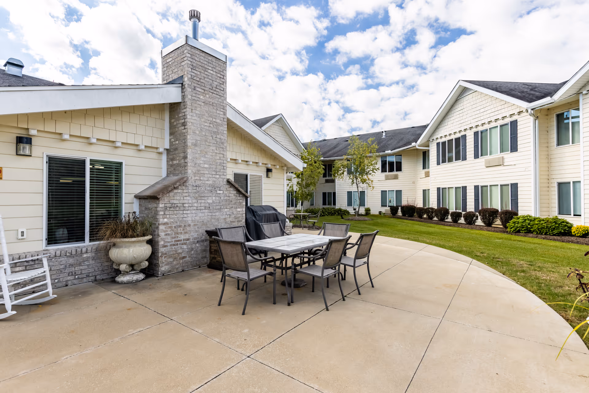 Outdoor patio area at Mulberry Gardens Senior Living featuring a brick fireplace, a dining table with six chairs, a white rocking chair, and a well-maintained lawn with shrubs and trees. The building exterior is light-colored with multiple windows under a partly cloudy sky.