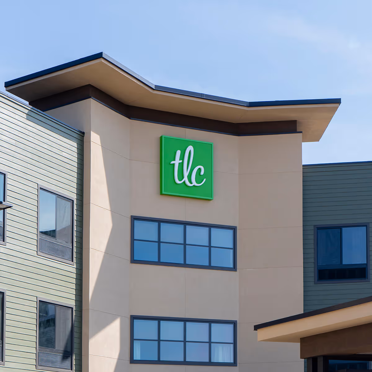 Exterior view of a modern building with beige and green walls, featuring a green sign with white lowercase letters 'tlc' mounted near the roof. The building has multiple windows and a clear blue sky in the background.