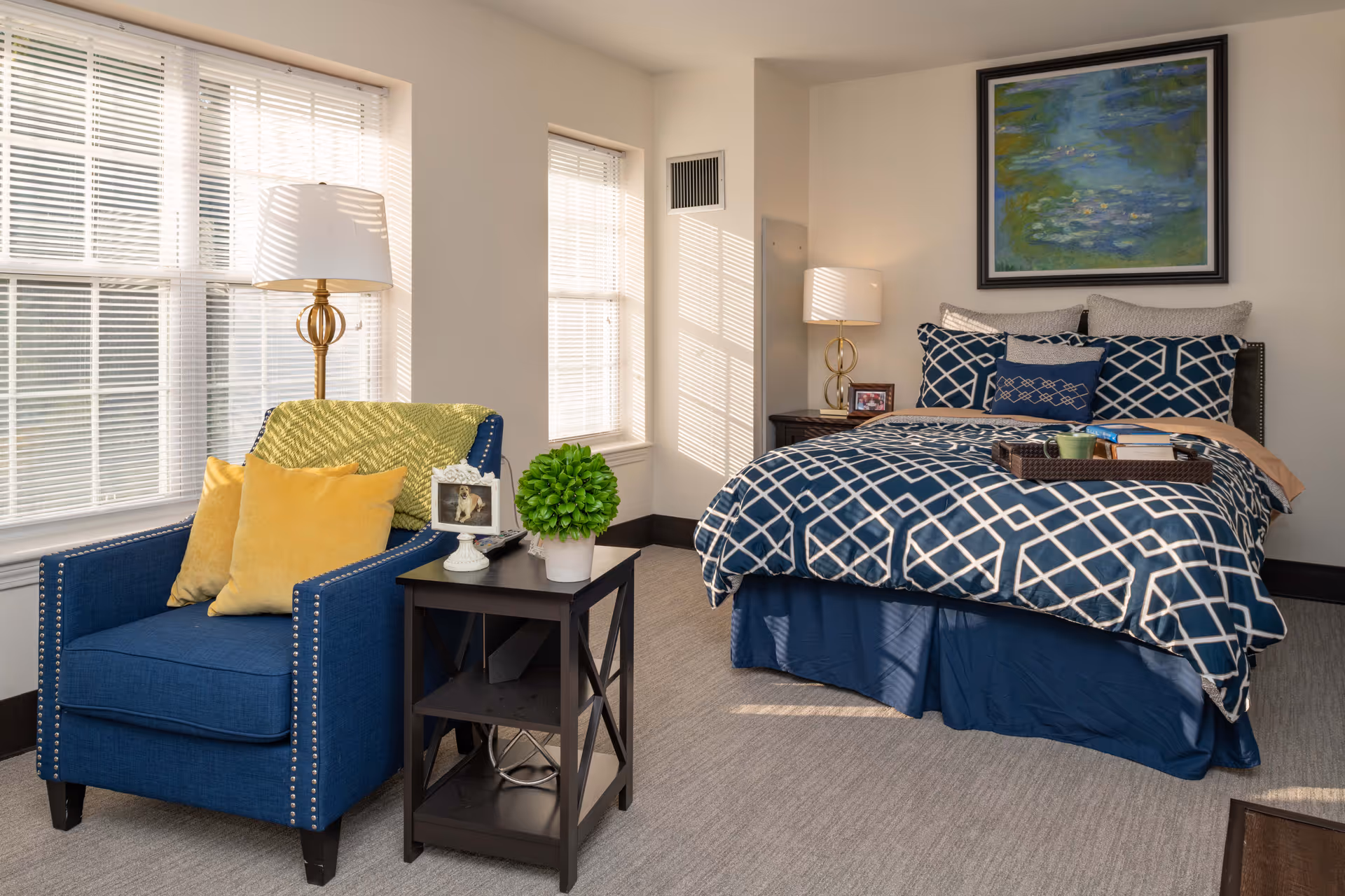 A bright bedroom with a large window letting in natural light. The room features a blue armchair with yellow pillows and a green throw blanket, a small black side table with a potted plant and a framed photo, and a bed with blue and white patterned bedding. There are two bedside tables with lamps, and a framed painting hangs on the wall above the bed.