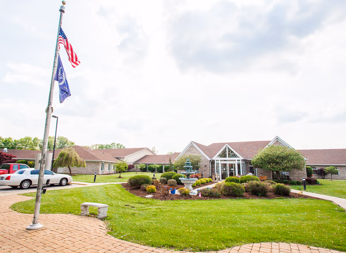 Front exterior of a single-story brick senior living facility with a landscaped lawn, fountain, flagpoles, and main entrance.