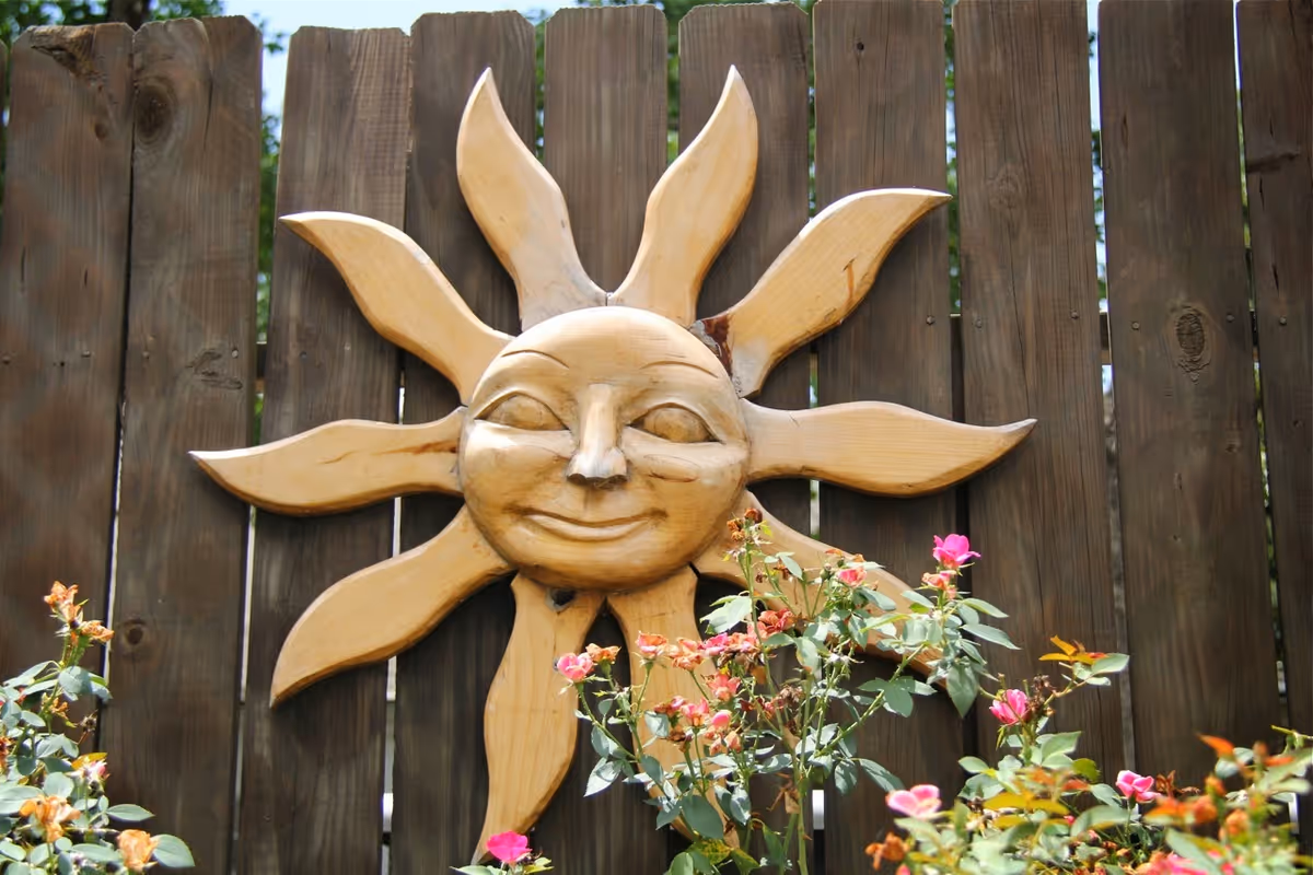 Wooden sun-face wall decoration mounted on a wooden fence above flowering plants.