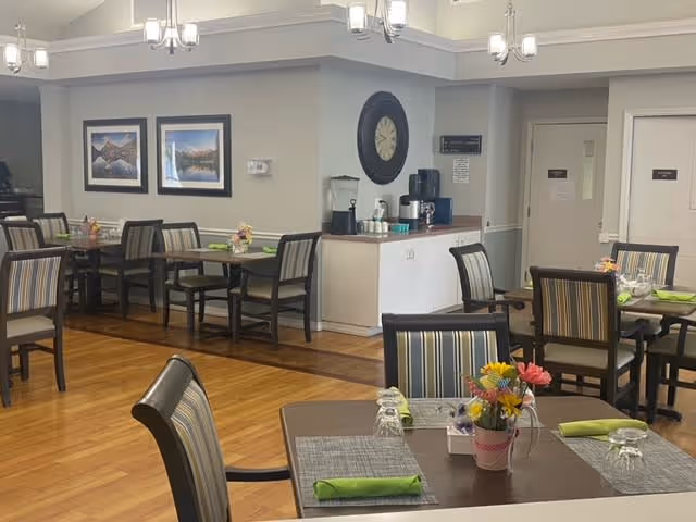 A dining room in a senior living facility with several tables and chairs arranged neatly. Each table has a placemat, a green napkin, upside-down glasses, and a small flower arrangement. The room has wooden flooring, light-colored walls, framed landscape pictures, and a large wall clock. A beverage station with a coffee maker and cups is visible in the background.