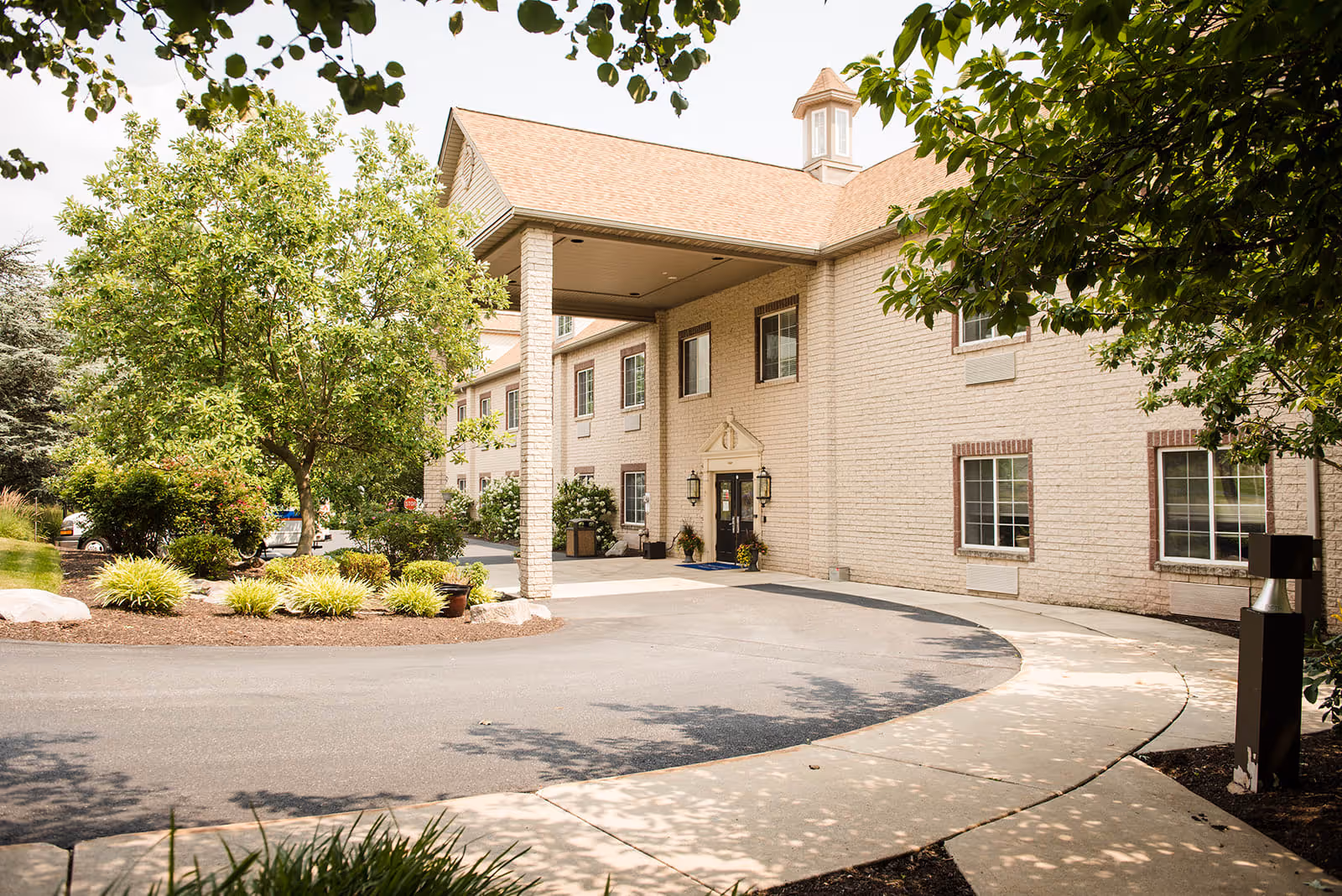 Exterior view of a senior living facility building with a covered entrance, surrounded by landscaped greenery including trees and shrubs, under a clear sky.