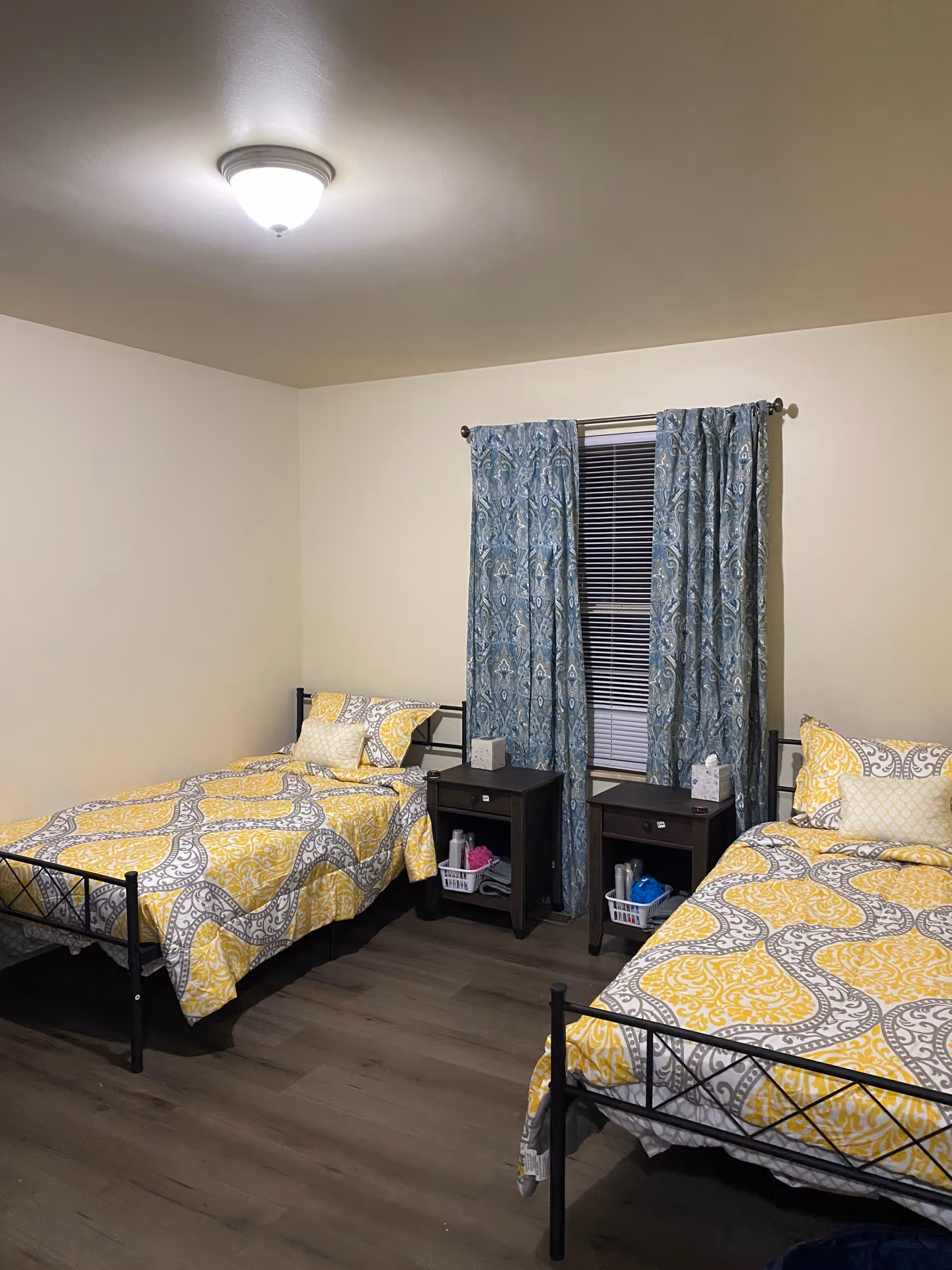 A bedroom with two twin beds featuring yellow and gray patterned bedding. Between the beds are two dark wooden nightstands with tissue boxes and small baskets containing personal care items. A window with blue patterned curtains and closed blinds is centered on the wall behind the nightstands. The room has light-colored walls and wood flooring, with a ceiling light fixture.